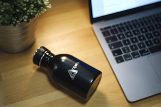 A sleek, black metal water bottle with a logo is placed on a wooden table beside an open laptop with a visible keyboard. A potted plant with green leaves is positioned to the left, adding a touch of greenery to the workspace.