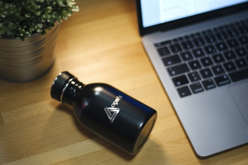 A sleek, black metal water bottle with a logo is placed on a wooden table beside an open laptop with a visible keyboard. A potted plant with green leaves is positioned to the left, adding a touch of greenery to the workspace.
