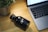 A sleek, black metal water bottle with a logo is placed on a wooden table beside an open laptop with a visible keyboard. A potted plant with green leaves is positioned to the left, adding a touch of greenery to the workspace.