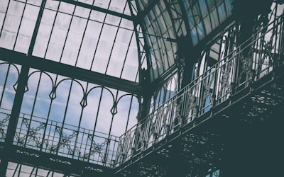 Intricate metal and glass architecture featuring tall, arched windows and ornate railings, allowing natural light to filter through. The structure exhibits a blend of classical and industrial design elements.