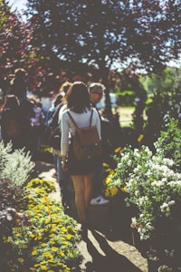 A group of people walking through a garden path lined with vibrant yellow and white flowers. The sunlight filters through the trees, casting dappled shadows on the ground. A woman in a white shirt and dark skirt is in the foreground, walking away from the camera, carrying a brown backpack. Others stroll along the path, surrounded by lush greenery.