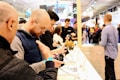 A group of people is gathered at a technology exhibition or store, engaging with electronic devices displayed on a white counter. The individuals appear focused on handling and examining these gadgets, suggesting an interactive and explorative environment. The background is busy with more people, likely attendees or visitors, moving around a well-lit space indicating a bustling atmosphere.