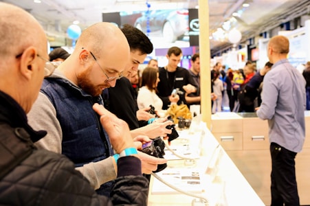 A group of people is gathered at a technology exhibition or store, engaging with electronic devices displayed on a white counter. The individuals appear focused on handling and examining these gadgets, suggesting an interactive and explorative environment. The background is busy with more people, likely attendees or visitors, moving around a well-lit space indicating a bustling atmosphere.