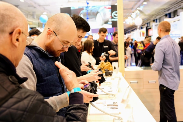 A group of people is gathered at a technology exhibition or store, engaging with electronic devices displayed on a white counter. The individuals appear focused on handling and examining these gadgets, suggesting an interactive and explorative environment. The background is busy with more people, likely attendees or visitors, moving around a well-lit space indicating a bustling atmosphere.