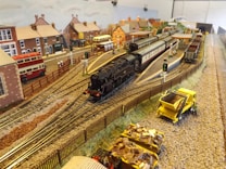 A detailed miniature railway model depicting a train station with black steam locomotives and carriages on tracks. Surrounding the tracks are model buildings resembling a small town with houses, buses, and a wooden fence. Additional elements include small figures, a yellow maintenance truck, and cargo wagons loaded with materials. The setting suggests a busy transport hub in a quaint village atmosphere.