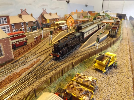 A detailed miniature railway model depicting a train station with black steam locomotives and carriages on tracks. Surrounding the tracks are model buildings resembling a small town with houses, buses, and a wooden fence. Additional elements include small figures, a yellow maintenance truck, and cargo wagons loaded with materials. The setting suggests a busy transport hub in a quaint village atmosphere.