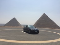 A black car is parked in a circular driveway in front of two large pyramids under a clear sky. The landscape is arid and expansive, with the pyramids dominating the background.