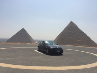 A black car is parked in a circular driveway in front of two large pyramids under a clear sky. The landscape is arid and expansive, with the pyramids dominating the background.