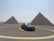 A black car is parked in a circular driveway in front of two large pyramids under a clear sky. The landscape is arid and expansive, with the pyramids dominating the background.