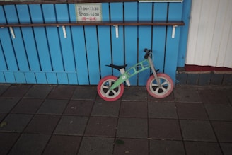 A small, colorfully designed balance bike with red wheels is leaned against a bright blue, wooden paneled wall. Above the bike is a shelf, and a sign displaying time schedules written in Japanese is attached to the wall. The ground is tiled with dark gray paving stones, some of which have fallen leaves.