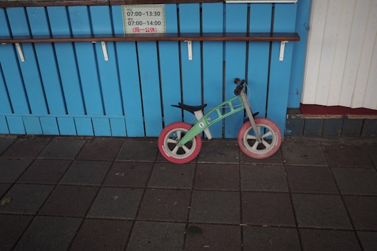 A small, colorfully designed balance bike with red wheels is leaned against a bright blue, wooden paneled wall. Above the bike is a shelf, and a sign displaying time schedules written in Japanese is attached to the wall. The ground is tiled with dark gray paving stones, some of which have fallen leaves.