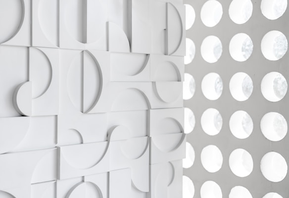 A geometric design featuring a pattern of raised semicircles and squares on a white textured wall next to a grid of circular cut-outs on an adjoining white surface.
