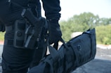 A person in black tactical gear is holding a large black duffel bag. They are equipped with a handgun holstered on their waist. The background consists of blurred greenery, suggesting an outdoor setting.