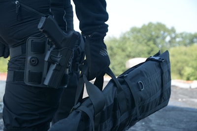 A person in black tactical gear is holding a large black duffel bag. They are equipped with a handgun holstered on their waist. The background consists of blurred greenery, suggesting an outdoor setting.
