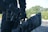 A person in black tactical gear is holding a large black duffel bag. They are equipped with a handgun holstered on their waist. The background consists of blurred greenery, suggesting an outdoor setting.