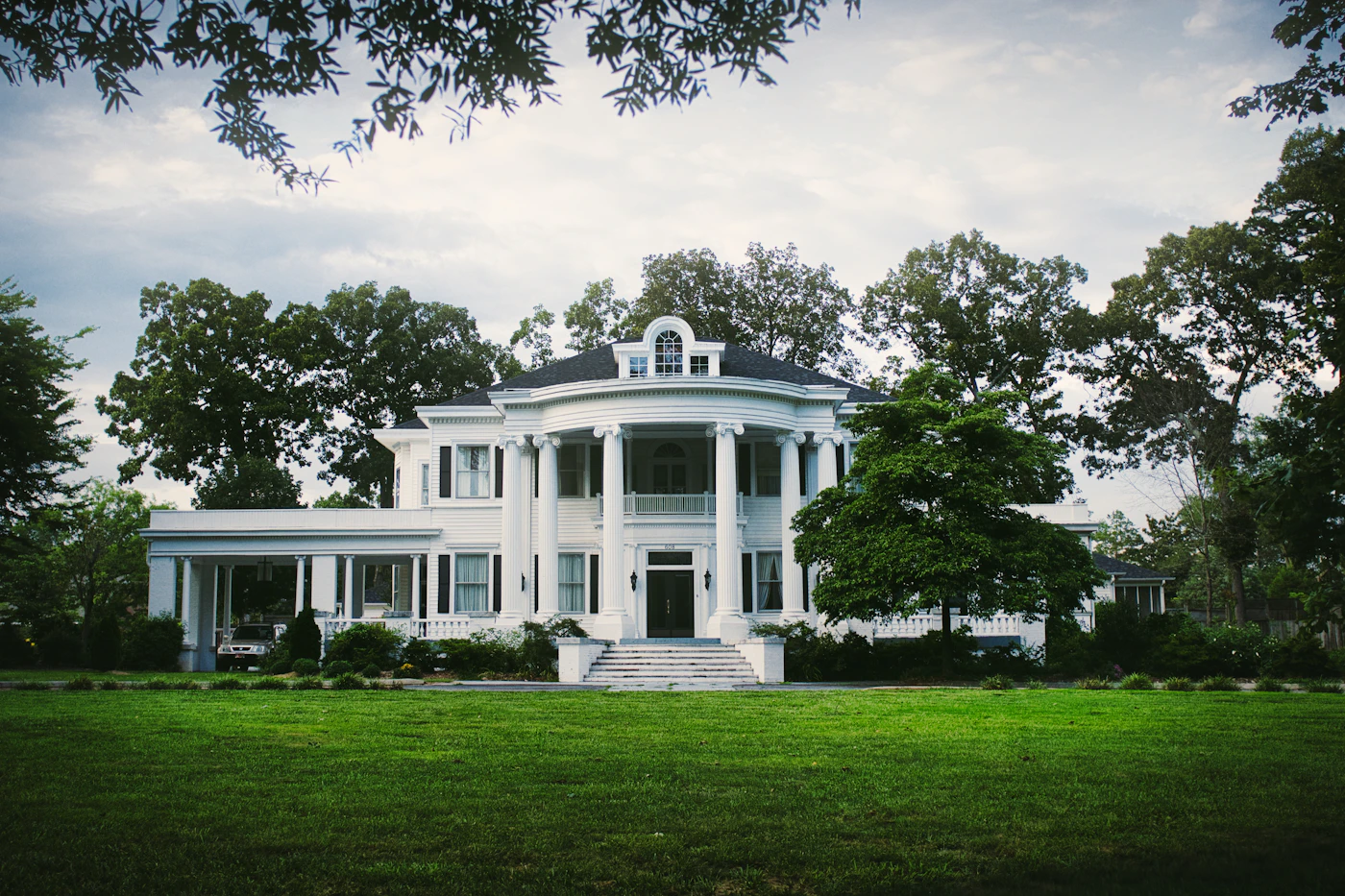 A white neoclassical country house with columns and wide front steps set in green parkland with mature trees surrounding the grounds