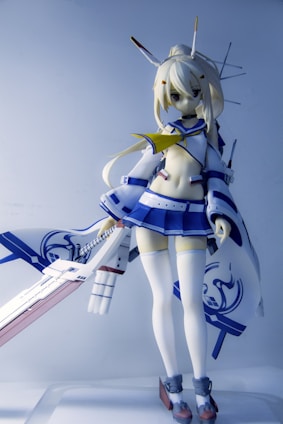 A detailed figure of an anime character with blonde hair is portrayed wearing a stylized outfit consisting of a blue and white sailor-style top and skirt, along with thigh-high stockings. The character is holding a large, ornate sword with intricate details. Antenna-like structures are part of the character's headgear, contributing to a futuristic or robotic design.