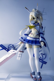 A detailed figure of an anime character with blonde hair is portrayed wearing a stylized outfit consisting of a blue and white sailor-style top and skirt, along with thigh-high stockings. The character is holding a large, ornate sword with intricate details. Antenna-like structures are part of the character's headgear, contributing to a futuristic or robotic design.