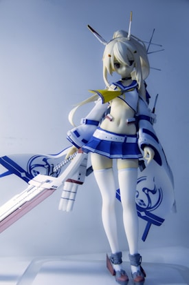 A detailed figure of an anime character with blonde hair is portrayed wearing a stylized outfit consisting of a blue and white sailor-style top and skirt, along with thigh-high stockings. The character is holding a large, ornate sword with intricate details. Antenna-like structures are part of the character's headgear, contributing to a futuristic or robotic design.