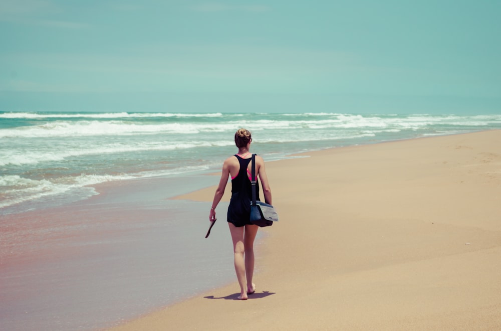 Frau in schwarzem Tanktop mit Tasche, die an der Küste spazieren geht