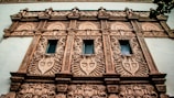 A detailed facade of a building featuring ornate architectural designs. The facade includes multiple decorative elements such as intricate patterns and motifs, with three vertical windows surrounded by lavish embellishments. The structure displays an elaborate style with intricate carvings and ornamental details, likely indicative of historical or classical architecture.