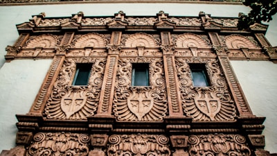 A detailed facade of a building featuring ornate architectural designs. The facade includes multiple decorative elements such as intricate patterns and motifs, with three vertical windows surrounded by lavish embellishments. The structure displays an elaborate style with intricate carvings and ornamental details, likely indicative of historical or classical architecture.