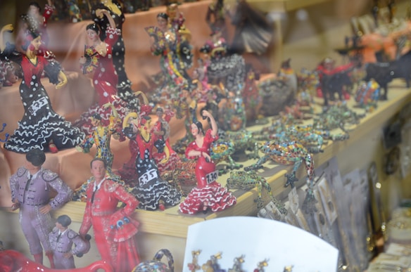A collection of colorful, intricately designed figurines on display in a shop. The figurines include dancers in traditional Spanish attire, such as flamenco dresses with polka dots. There are also other animal figurines like frogs displayed alongside them. The scene is vibrant with a variety of poses indicating movement and celebration.