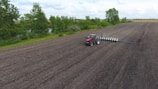 A red tractor with a planting attachment drives across a large, freshly tilled agricultural field. The field is expansive, bordered by a line of green trees and a small water body on one side. The sky is cloudy, providing a peaceful backdrop to the rural landscape.