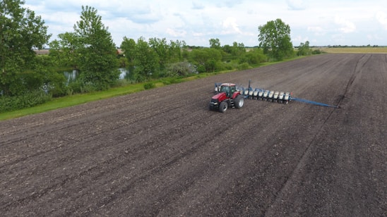 A red tractor with a planting attachment drives across a large, freshly tilled agricultural field. The field is expansive, bordered by a line of green trees and a small water body on one side. The sky is cloudy, providing a peaceful backdrop to the rural landscape.