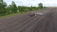 A red tractor with a planting attachment drives across a large, freshly tilled agricultural field. The field is expansive, bordered by a line of green trees and a small water body on one side. The sky is cloudy, providing a peaceful backdrop to the rural landscape.