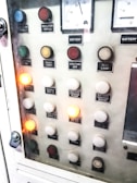 A control panel with various labeled buttons and switches, including color-coded round indicators. Labels such as insulation test, night light, lighting on, battery charge float, and failure notices are present. Some buttons are lit, indicating active status, while others remain unlit. A voltmeter gauge is visible at the top, showing voltage levels.