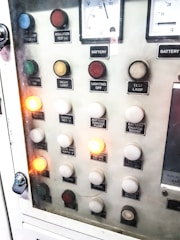A control panel with various labeled buttons and switches, including color-coded round indicators. Labels such as insulation test, night light, lighting on, battery charge float, and failure notices are present. Some buttons are lit, indicating active status, while others remain unlit. A voltmeter gauge is visible at the top, showing voltage levels.