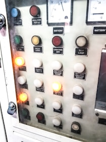 A control panel with various labeled buttons and switches, including color-coded round indicators. Labels such as insulation test, night light, lighting on, battery charge float, and failure notices are present. Some buttons are lit, indicating active status, while others remain unlit. A voltmeter gauge is visible at the top, showing voltage levels.