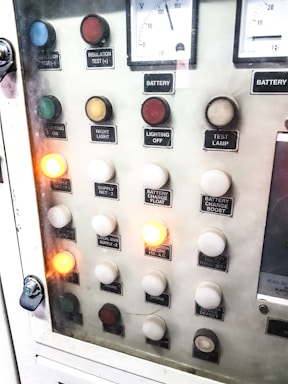 A control panel with various labeled buttons and switches, including color-coded round indicators. Labels such as insulation test, night light, lighting on, battery charge float, and failure notices are present. Some buttons are lit, indicating active status, while others remain unlit. A voltmeter gauge is visible at the top, showing voltage levels.