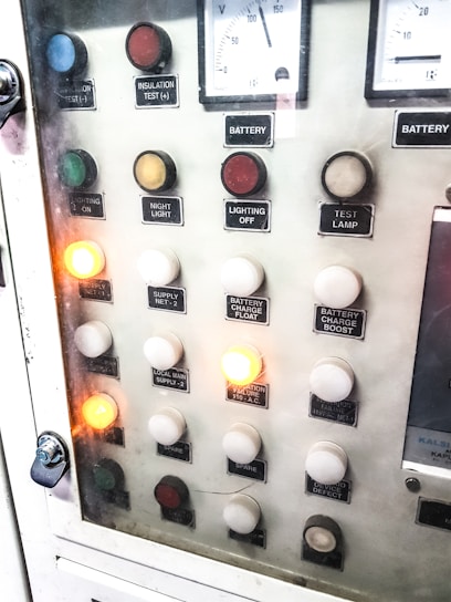 A control panel with various labeled buttons and switches, including color-coded round indicators. Labels such as insulation test, night light, lighting on, battery charge float, and failure notices are present. Some buttons are lit, indicating active status, while others remain unlit. A voltmeter gauge is visible at the top, showing voltage levels.