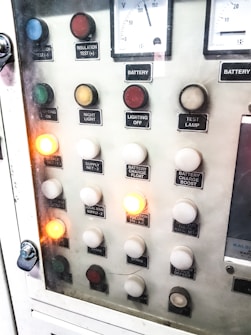 A control panel with various labeled buttons and switches, including color-coded round indicators. Labels such as insulation test, night light, lighting on, battery charge float, and failure notices are present. Some buttons are lit, indicating active status, while others remain unlit. A voltmeter gauge is visible at the top, showing voltage levels.