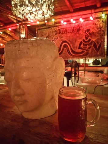 A serene Buddha head sculpture sits on a wooden table beside a frothy glass of beer. In the background, a decorative chandelier made of circular elements hangs from the ceiling, and colorful lights illuminate a mural featuring artistic designs and a partially visible word 'SHAMANIA'. The setting gives a rustic and relaxed ambiance.