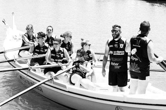 A group of people are rowing in a boat on the water. They appear to be part of a team, as they are wearing matching sleeveless shirts with logos and text. The image is in black and white, showing them in action under a bright setting.