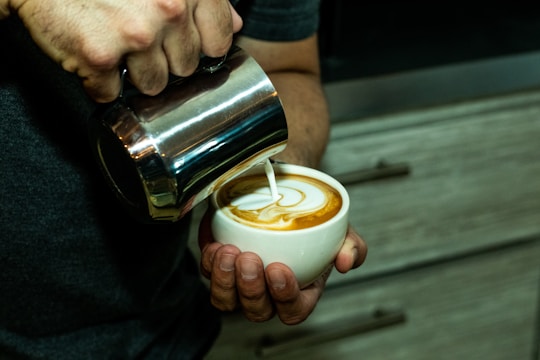 A person is skillfully pouring steamed milk into a cup of coffee, creating intricate latte art on the surface. The hands holding the cup and pitcher are visible, and the background consists of wooden drawers.