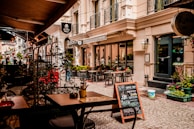 A quaint outdoor café setting on a cobblestone street with several empty tables and chairs under a canopy. The street is lined with a charming boutique hotel and shopfronts adorned with potted plants and decorative lanterns. A sidewalk chalkboard advertises coffee selections like espresso and latte.