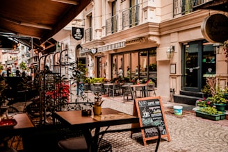 A quaint outdoor café setting on a cobblestone street with several empty tables and chairs under a canopy. The street is lined with a charming boutique hotel and shopfronts adorned with potted plants and decorative lanterns. A sidewalk chalkboard advertises coffee selections like espresso and latte.