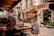 A quaint outdoor café setting on a cobblestone street with several empty tables and chairs under a canopy. The street is lined with a charming boutique hotel and shopfronts adorned with potted plants and decorative lanterns. A sidewalk chalkboard advertises coffee selections like espresso and latte.