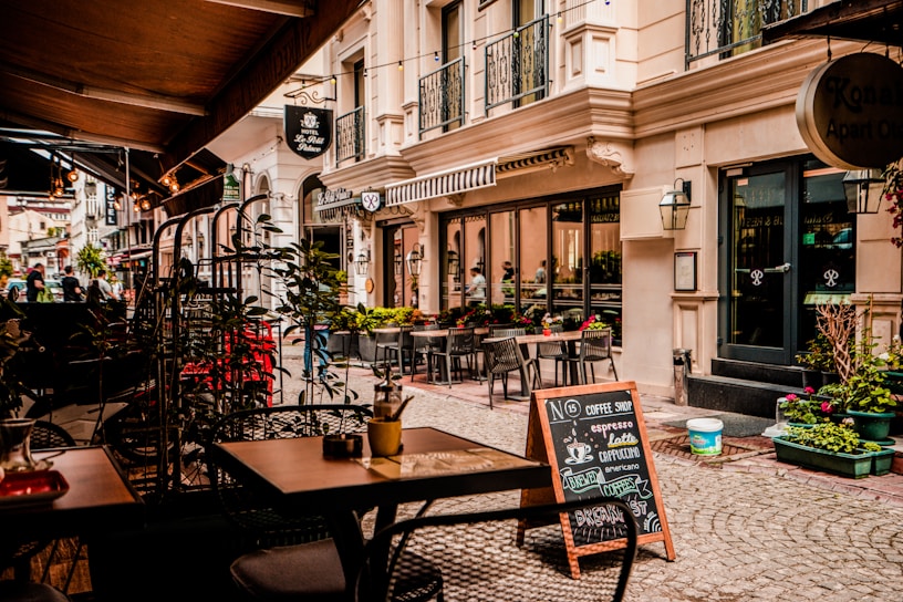 A quaint outdoor café setting on a cobblestone street with several empty tables and chairs under a canopy. The street is lined with a charming boutique hotel and shopfronts adorned with potted plants and decorative lanterns. A sidewalk chalkboard advertises coffee selections like espresso and latte.