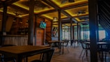 A spacious and elegantly designed restaurant interior with wooden furniture and warm lighting. The room features several tables and chairs, a wall-mounted TV, ceiling fans, and wooden beams. Large windows provide natural light and a view of the greenery outside.