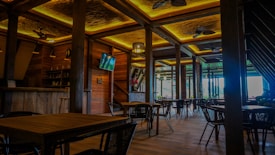 A spacious and elegantly designed restaurant interior with wooden furniture and warm lighting. The room features several tables and chairs, a wall-mounted TV, ceiling fans, and wooden beams. Large windows provide natural light and a view of the greenery outside.