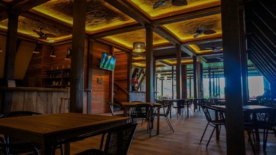 A spacious and elegantly designed restaurant interior with wooden furniture and warm lighting. The room features several tables and chairs, a wall-mounted TV, ceiling fans, and wooden beams. Large windows provide natural light and a view of the greenery outside.