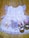 A delicate children's dress made of white tulle fabric adorned with silver sequins. It features ruffled sleeves and a floral embellishment near the neckline. Accompanying the dress are a set of gold bangles, a hair accessory with furry details and a bow, and a pair of gold sandals with pink pom-poms.