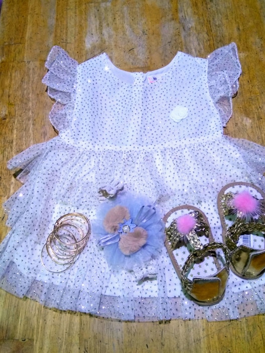 A delicate children's dress made of white tulle fabric adorned with silver sequins. It features ruffled sleeves and a floral embellishment near the neckline. Accompanying the dress are a set of gold bangles, a hair accessory with furry details and a bow, and a pair of gold sandals with pink pom-poms.