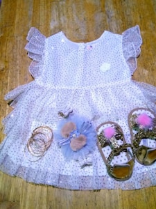 A delicate children's dress made of white tulle fabric adorned with silver sequins. It features ruffled sleeves and a floral embellishment near the neckline. Accompanying the dress are a set of gold bangles, a hair accessory with furry details and a bow, and a pair of gold sandals with pink pom-poms.