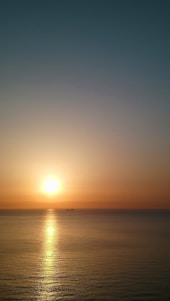 A calm ocean horizon at sunrise with a silhouette of a cargo ship in the distance.